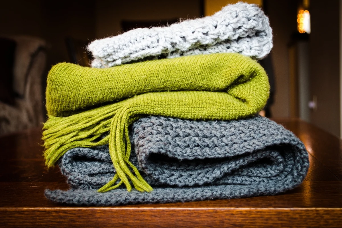 Folded knit layers stacked on a scratched oak table beside an open suitcase, packing cubes unzipped, a paper checklist with a pencil resting on the lid, late afternoon indoor light, 1400x933