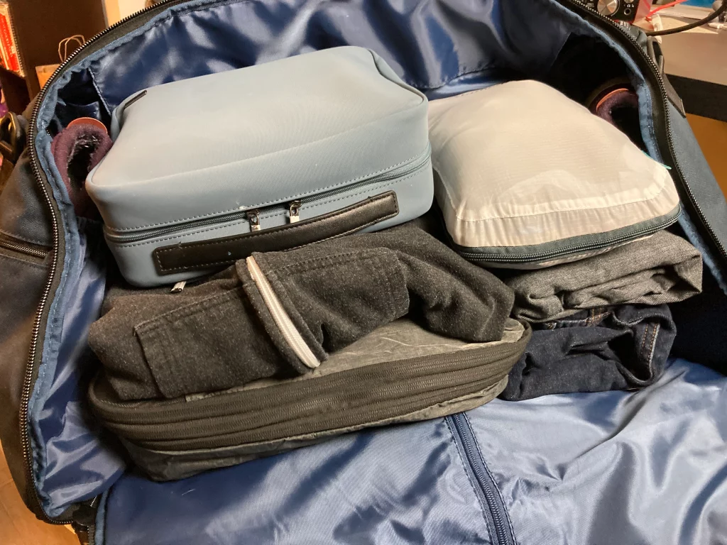 Half-packed duffel on a wooden floor beside a printed itinerary with circled connection times, two shirts still on hangers, a pair of walking shoes set apart from the bag, late indoor light, 1400x933
