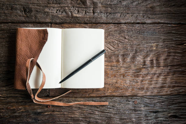 Worn studio desk in side window light, an open cloth notebook with a packing sketch, a coiled tape measure, and a printed ticket sleeve lying on oak grain, empty chair pushed back, 1920x1080