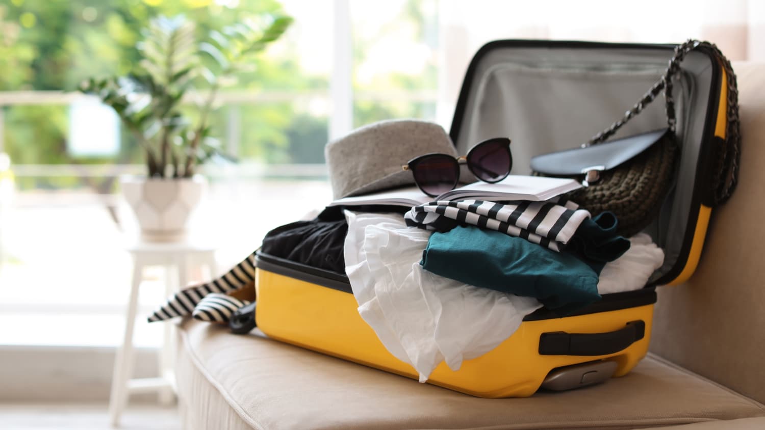 Open hard-shell suitcase on a rumpled linen bed in morning window light, folded knits stacked beside a canvas toiletry pouch, a cloth tape measure draped over the lid, pale plaster wall behind, 1920x1080
